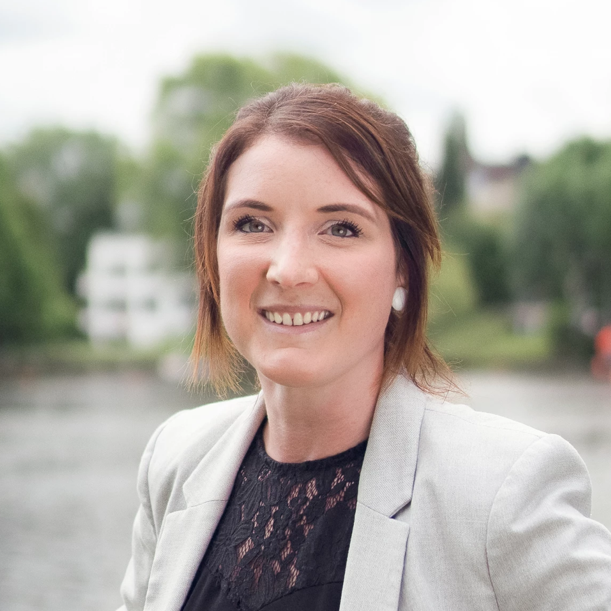 Eine Frau mit braunen Haaren in einem hellen Blazer lächelt vor einer unscharfen, natürlichen Wasserlandschaft.