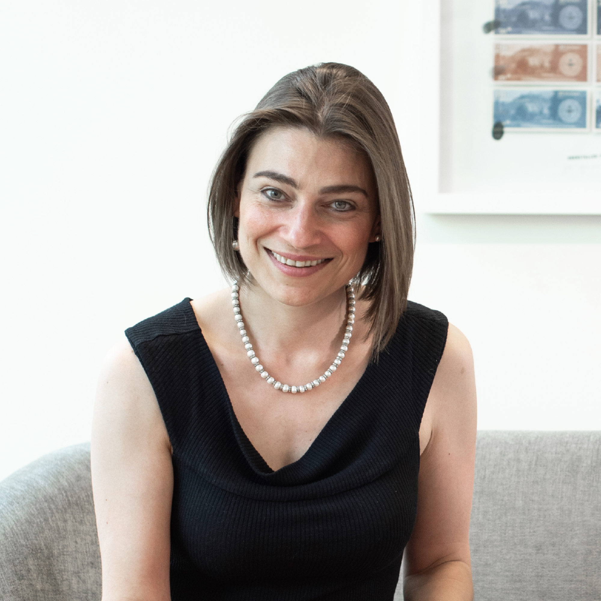 Eine Frau mit schulterlangen, braunen Haaren sitzt auf einem grauen Sofa und lächelt freundlich. Sie trägt ein schwarzes, ärmelloses Oberteil und eine Perlenkette. Im Hintergrund sind kunstvolle Grafiken oder Drucke an der Wand sichtbar.