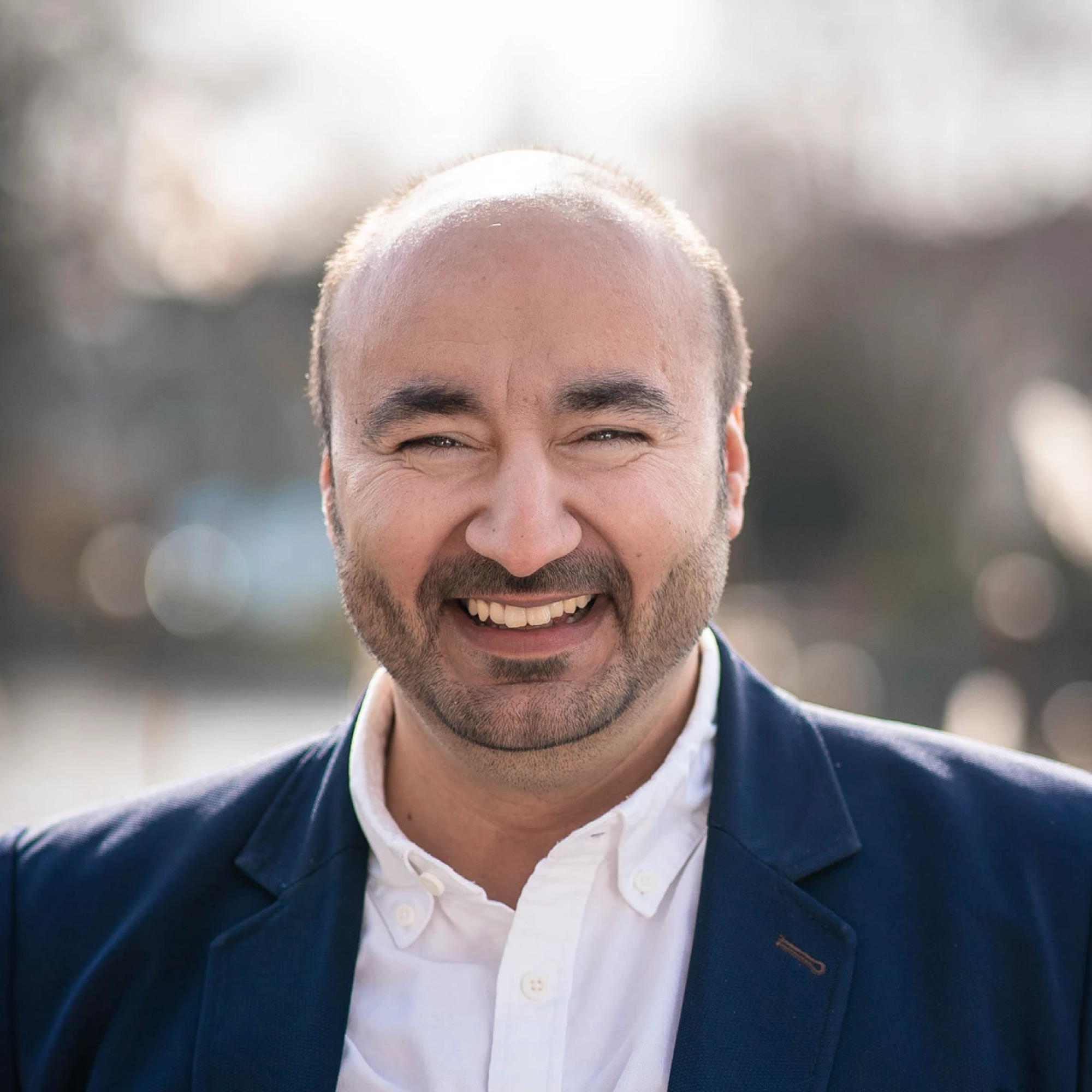 Ein Mann mit Glatze und Bart lächelt fröhlich in die Kamera, trägt ein dunkles Sakko und ein weißes Hemd, während unscharfe Lichtpunkte im Hintergrund zu sehen sind.