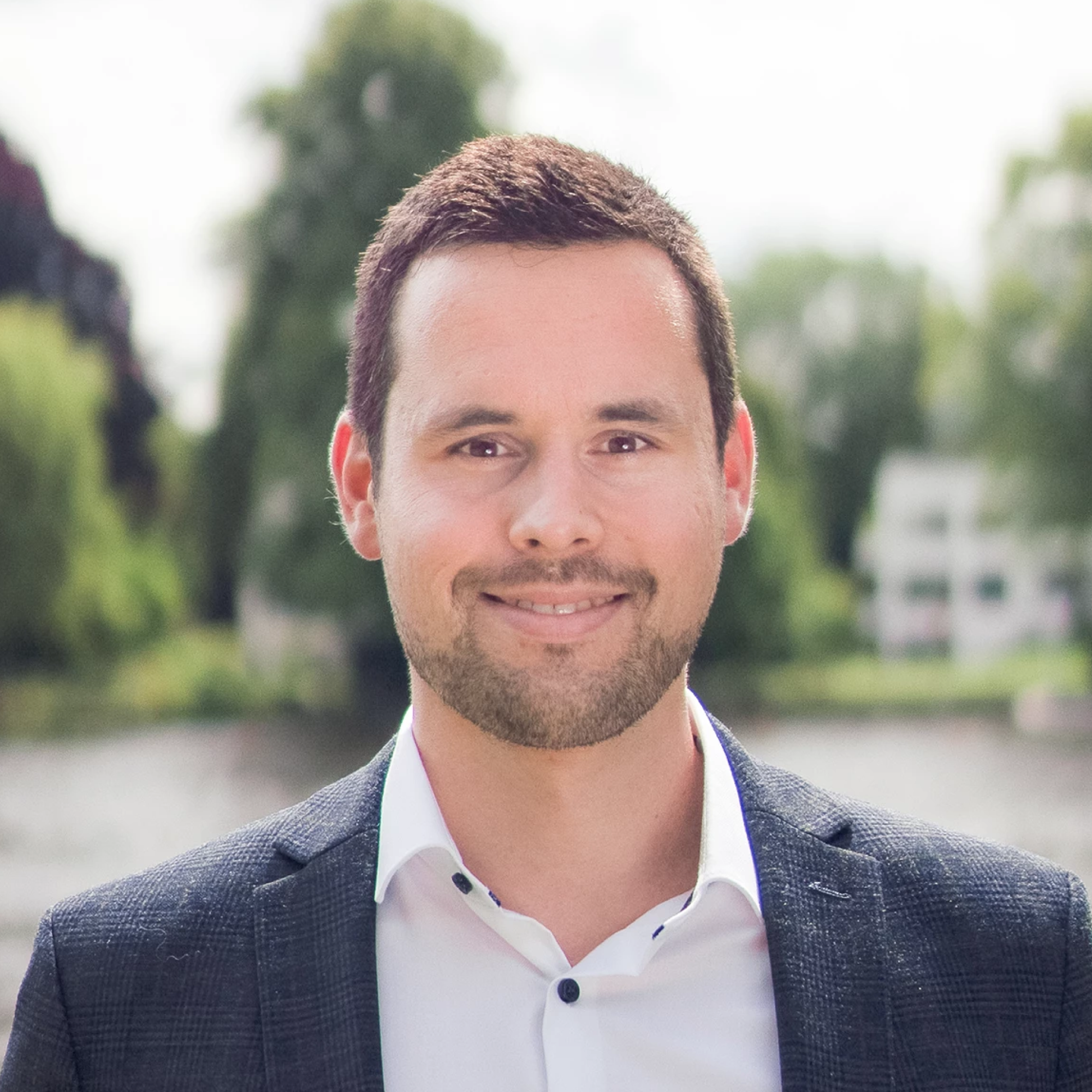 Ein Mann mit dunklem Haar und einem gepflegten Bart lächelt in die Kamera, während er ein dunkelgraues Jackett und ein weißes Hemd trägt. Im Hintergrund sind unscharfe Bäume und ein Gewässer zu sehen.
