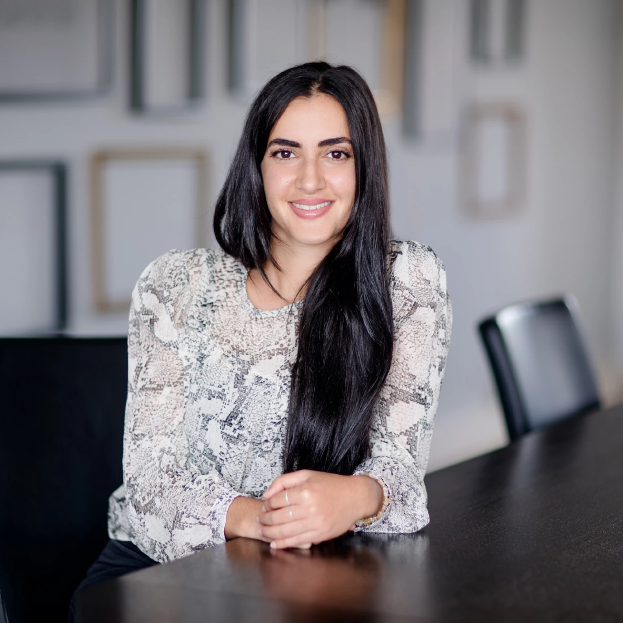 Eine lächelnde Frau mit langen, dunklen Haaren sitzt an einem Tisch und lehnt sich mit einem Arm leicht auf die Tischplatte. Sie trägt ein blusenartiges Oberteil mit Schlangenledermuster und ist in einem hellen, modernen Raum mit verschwommenen Bildrahmen im Hintergrund abgebildet.
