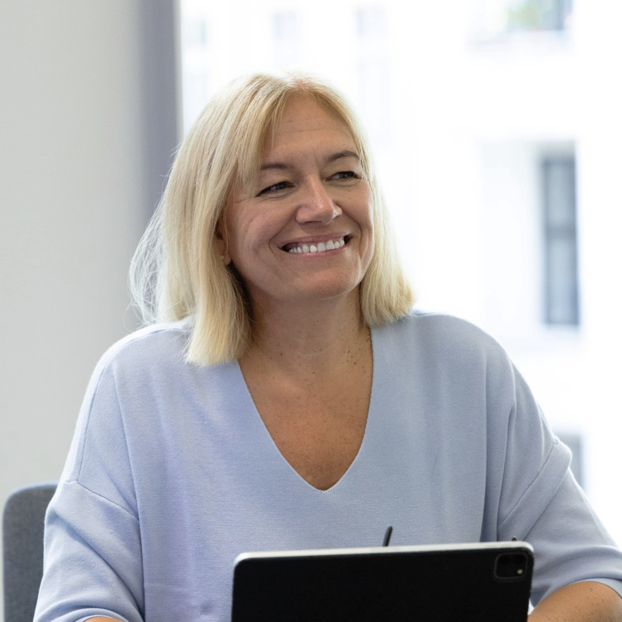 Eine blonde Frau mit einem freundlichen Lächeln sitzt in einem modernen Büro, während sie auf ein Tablet schaut. Die Umgebung ist hell und freundlich.