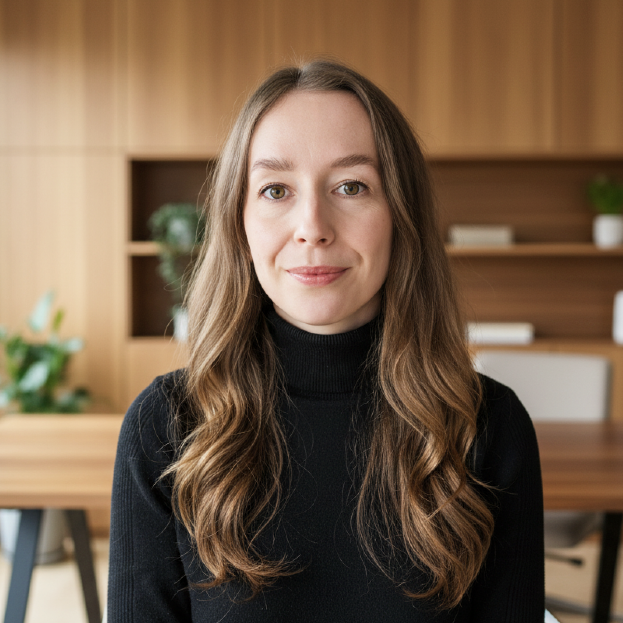 Eine Frau mit langen, gewellten braunen Haaren trägt einen schwarzen Rollkragenpullover und sitzt in einem modernen Büro mit holzverkleideten Wänden und Pflanzen im Hintergrund.