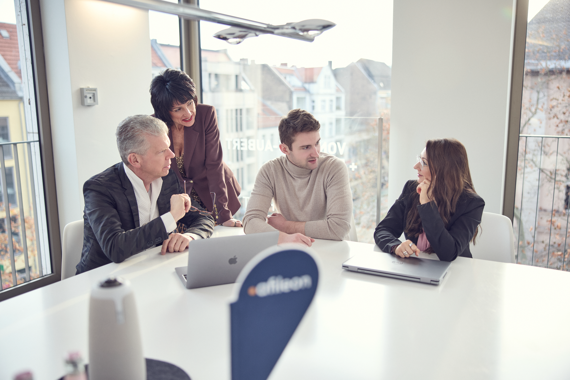 Drei Personen sitzen an einem Tisch und diskutieren, während eine weitere Person über ihnen steht und aufmerksam zuhört. Auf dem Tisch befinden sich Laptops und Dokumente. Im Hintergrund sind Fenster mit Blick auf eine städtische Landschaft zu sehen.