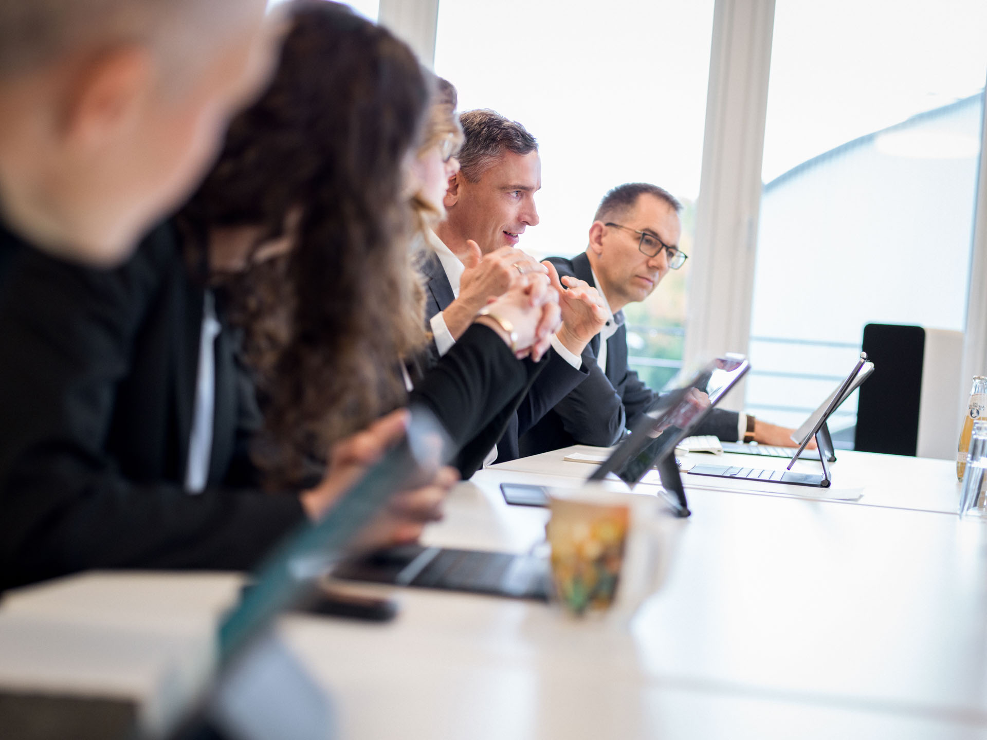 Eine Gruppe von Personen in formeller Kleidung sitzt an einem Konferenztisch, während sie aufmerksam einem Vortrag zuschauen. Auf dem Tisch befinden sich Tablets und eine Tasse. Im Hintergrund sind große Fenster zu sehen, die Tageslicht hereinlassen.