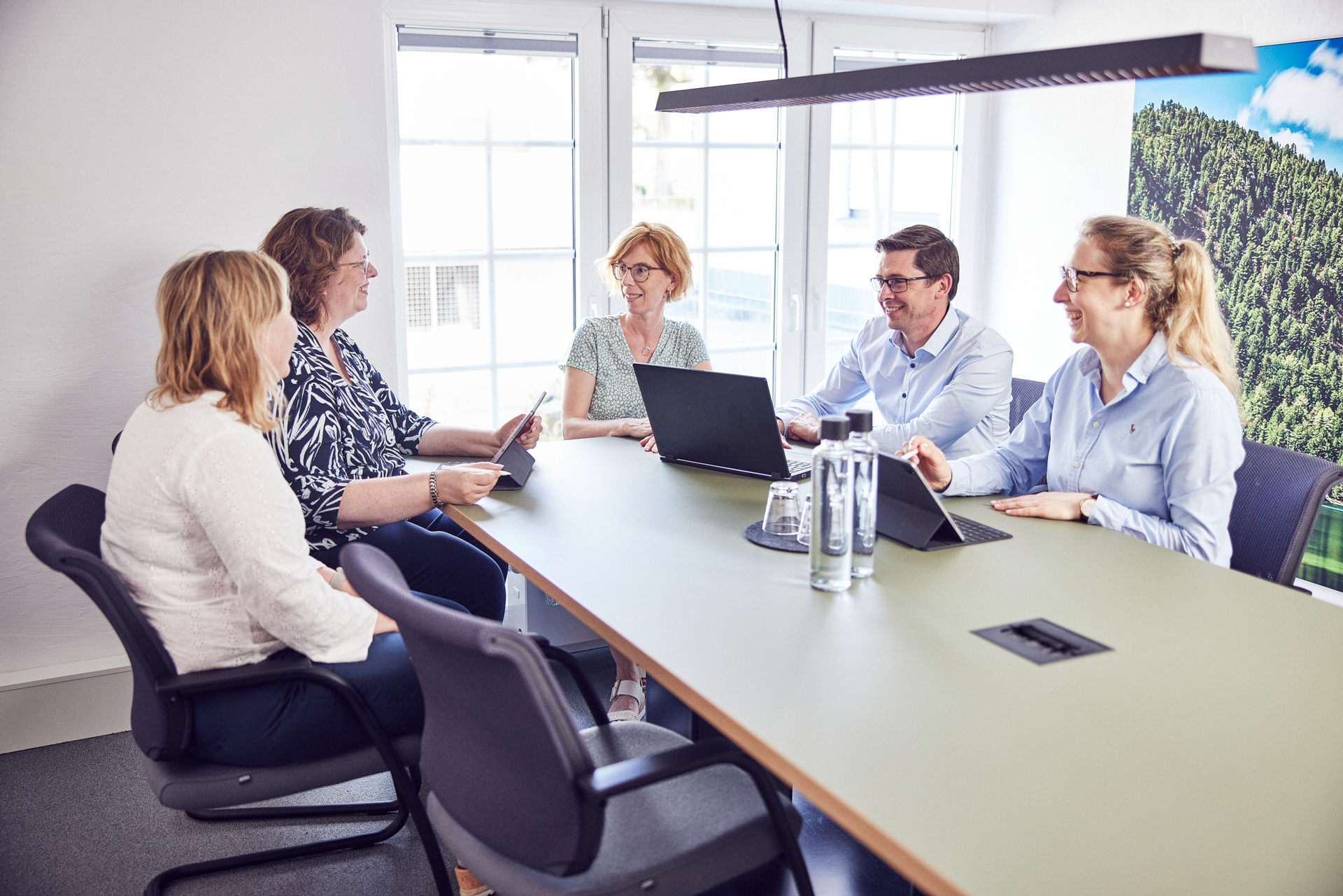 Fünf Personen sitzen um einen Tisch in einem modernen Besprechungsraum und diskutieren, während Laptops und Tablets auf dem Tisch liegen. Eine große Fensterfront lässt viel Tageslicht herein.