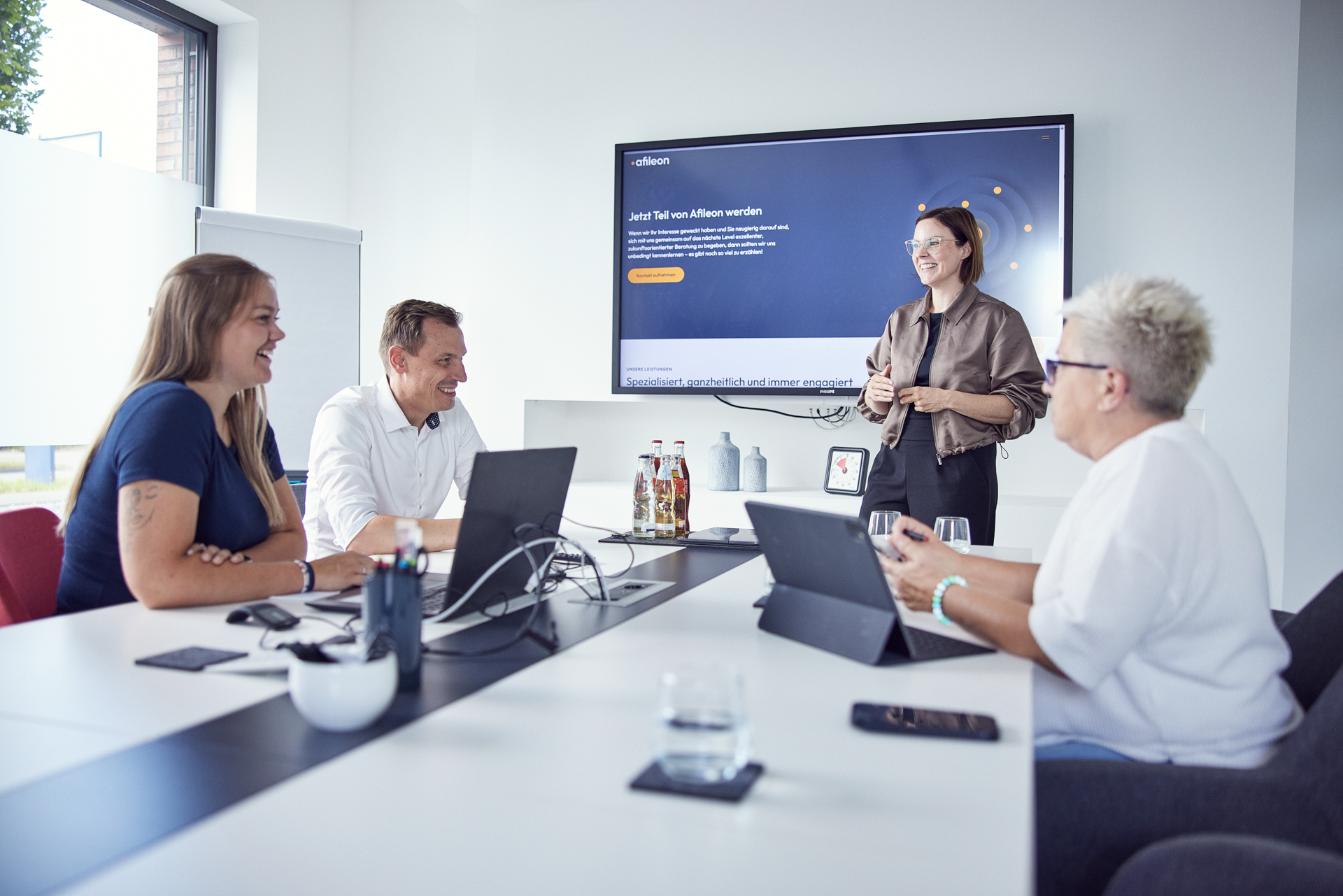 Eine Person präsentiert in einem modernen Konferenzraum vor einem großen Bildschirm, während drei weitere Personen am Tisch sitzen und aufmerksam zuhören. Auf dem Bildschirm steht der Text “Jetzt Teil von Affilion werden”.