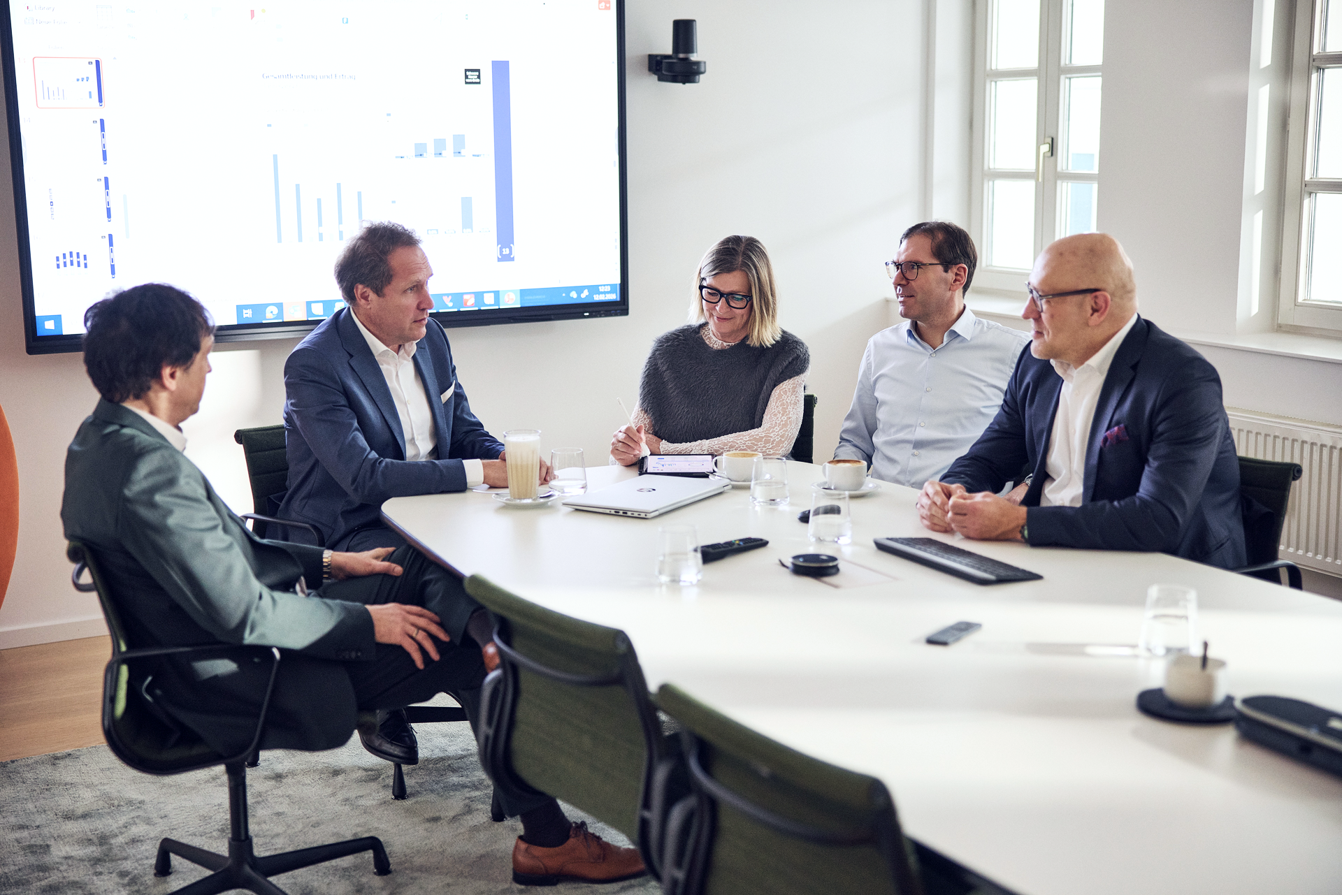 Eine Gruppe von fünf Personen sitzt an einem modernen Konferenztisch in einem hellen Besprechungsraum. Auf einem großen Bildschirm im Hintergrund sind Grafiken und Daten visualisiert. Alle Anwesenden sind mit Notizen und Getränken beschäftigt, während sie sich engagiert austauschen.