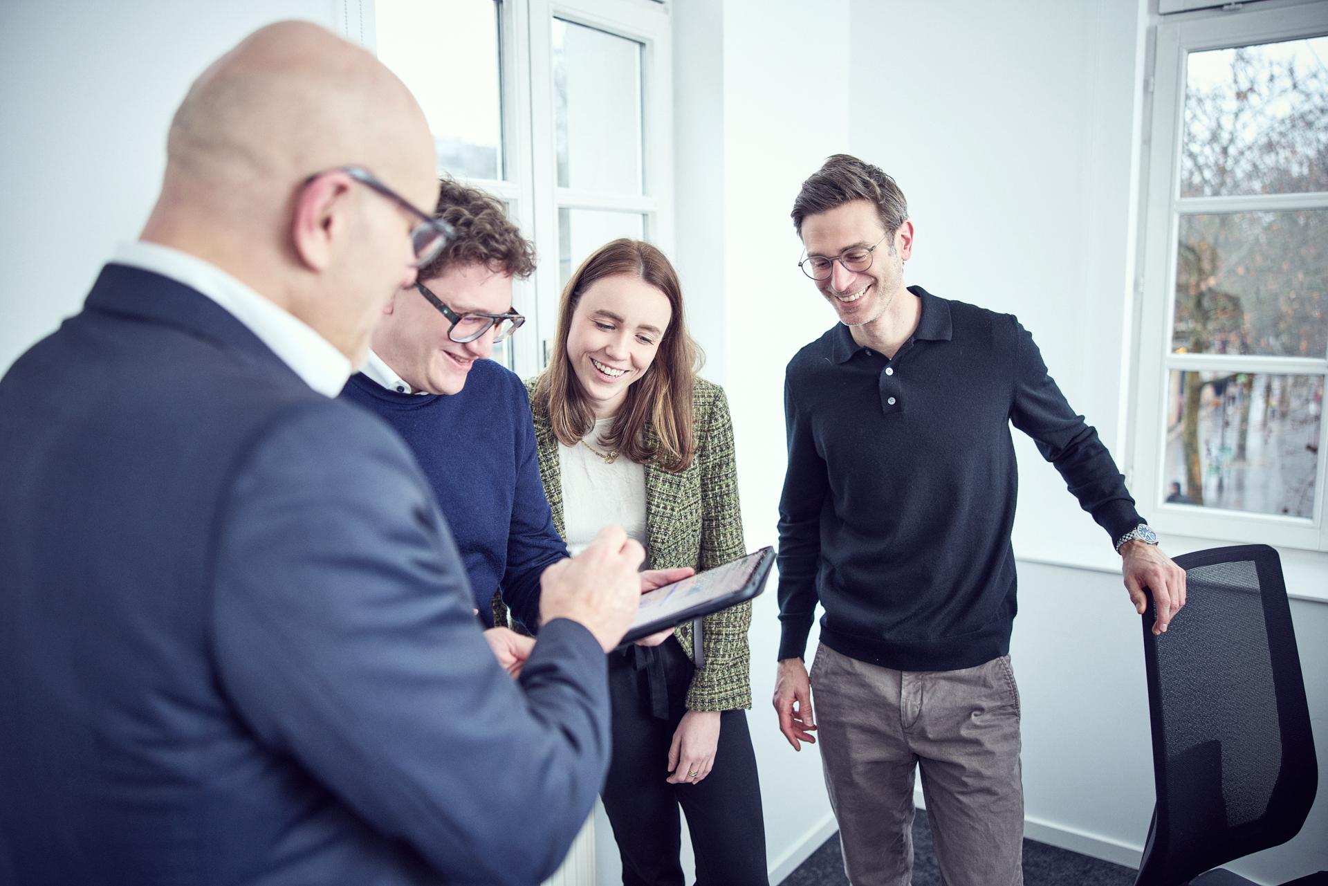 Vier Personen in einem hellen Büro schauen gemeinsam auf ein Tablet. Eine Person im Vordergrund zeigt etwas auf dem Bildschirm, während die anderen interessiert schauen und lächeln. Es herrscht eine freundliche und kooperative Atmosphäre.