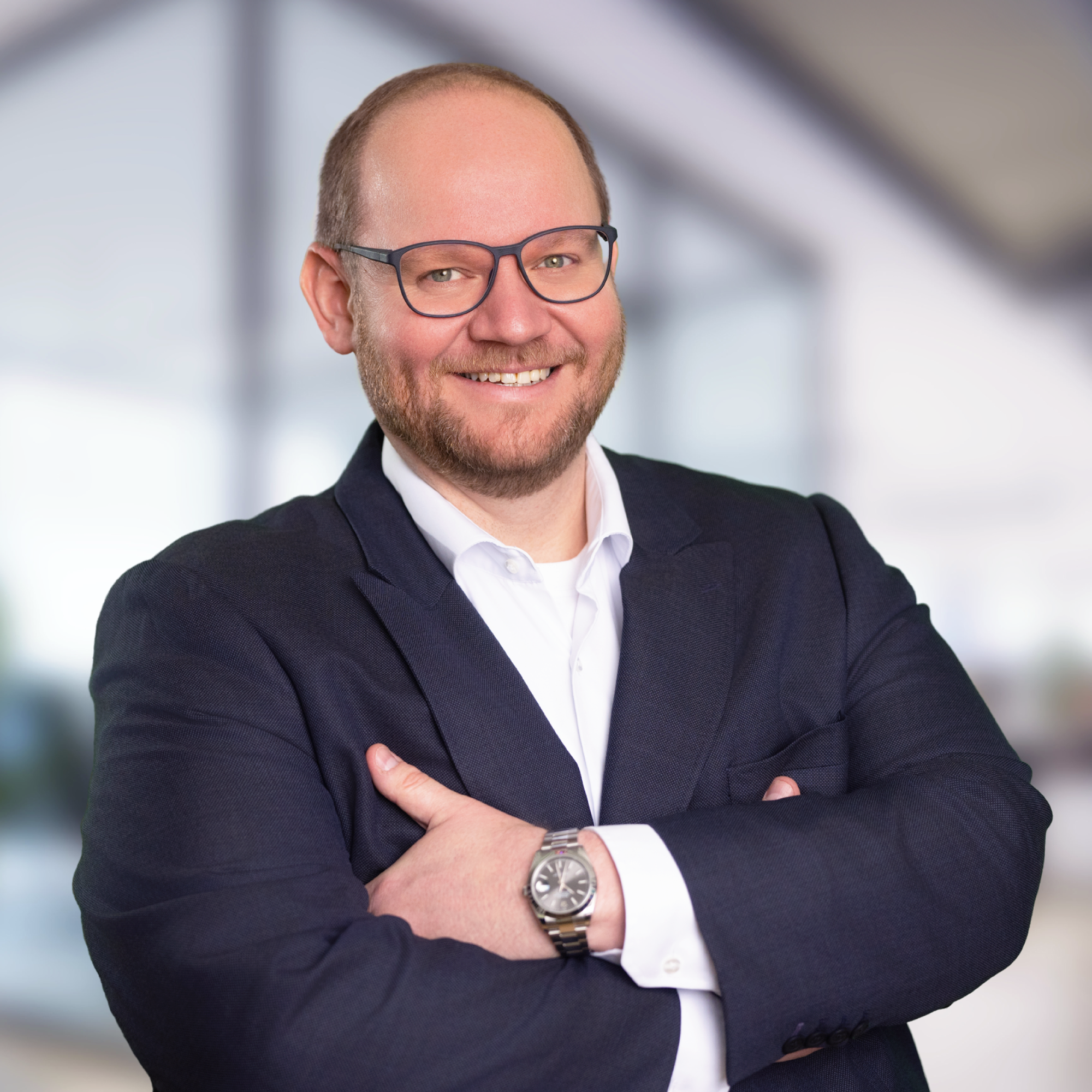 Ein lächelnder Mann mit Brille steht mit verschränkten Armen in formeller Kleidung vor einem modernen, hellen Hintergrund.
