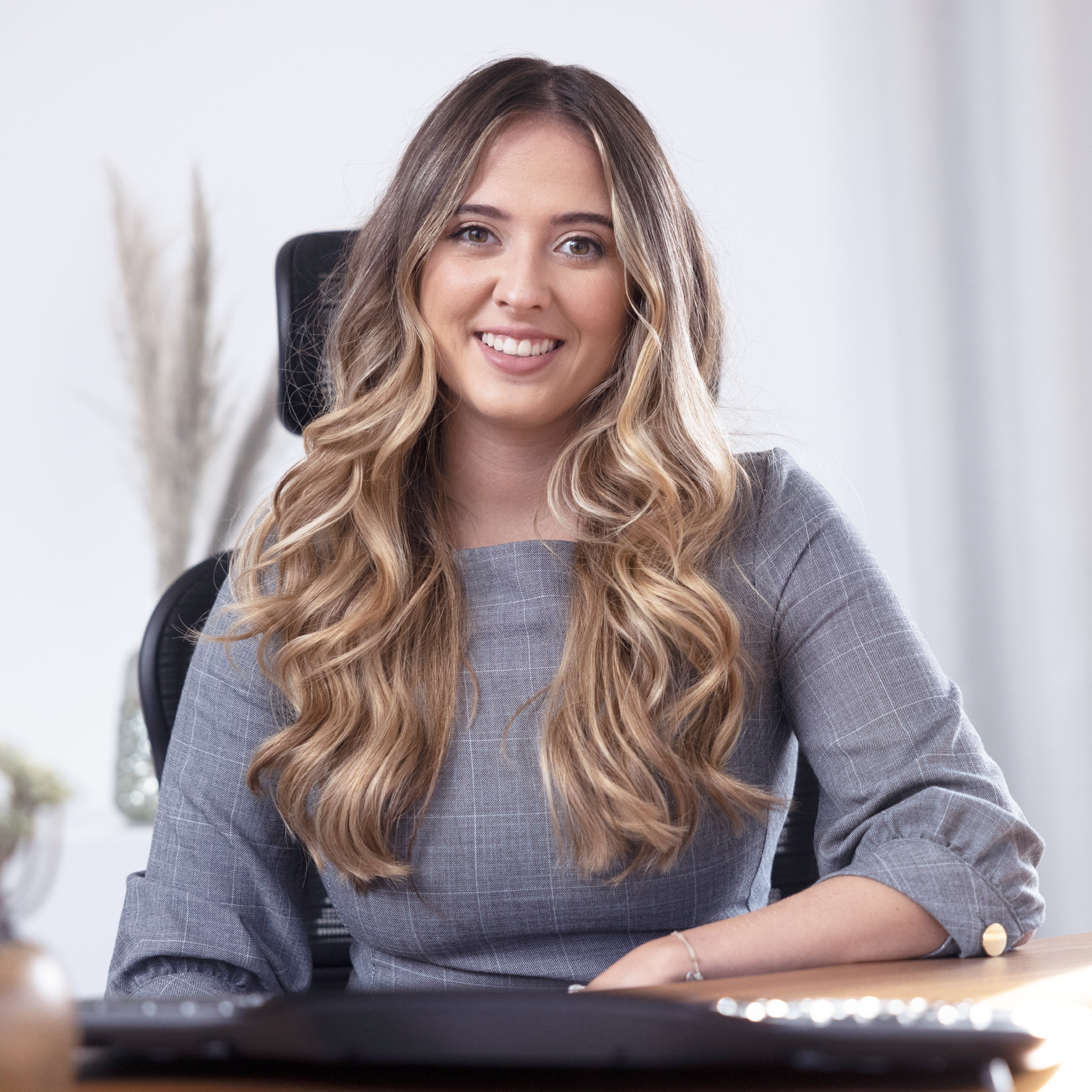 Eine junge Frau mit langen, welligen Haaren sitzt an einem Schreibtisch in einem modernen Büro. Sie trägt ein graues, elegantes Kleid und lächelt freundlich in die Kamera. Im Hintergrund sind Pflanzen und eine helle, einladende Umgebung zu sehen.
