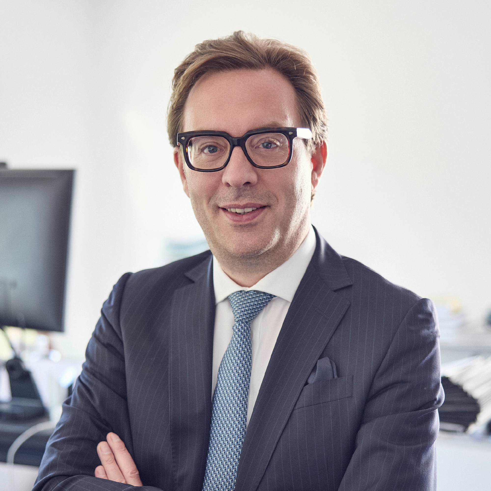 Ein lächelnder Mann in einem dunkelblauen Anzug mit einem gestreiften Hemd und einer hellblauen Krawatte steht mit verschränkten Armen vor einem modernen Bürohintergrund.