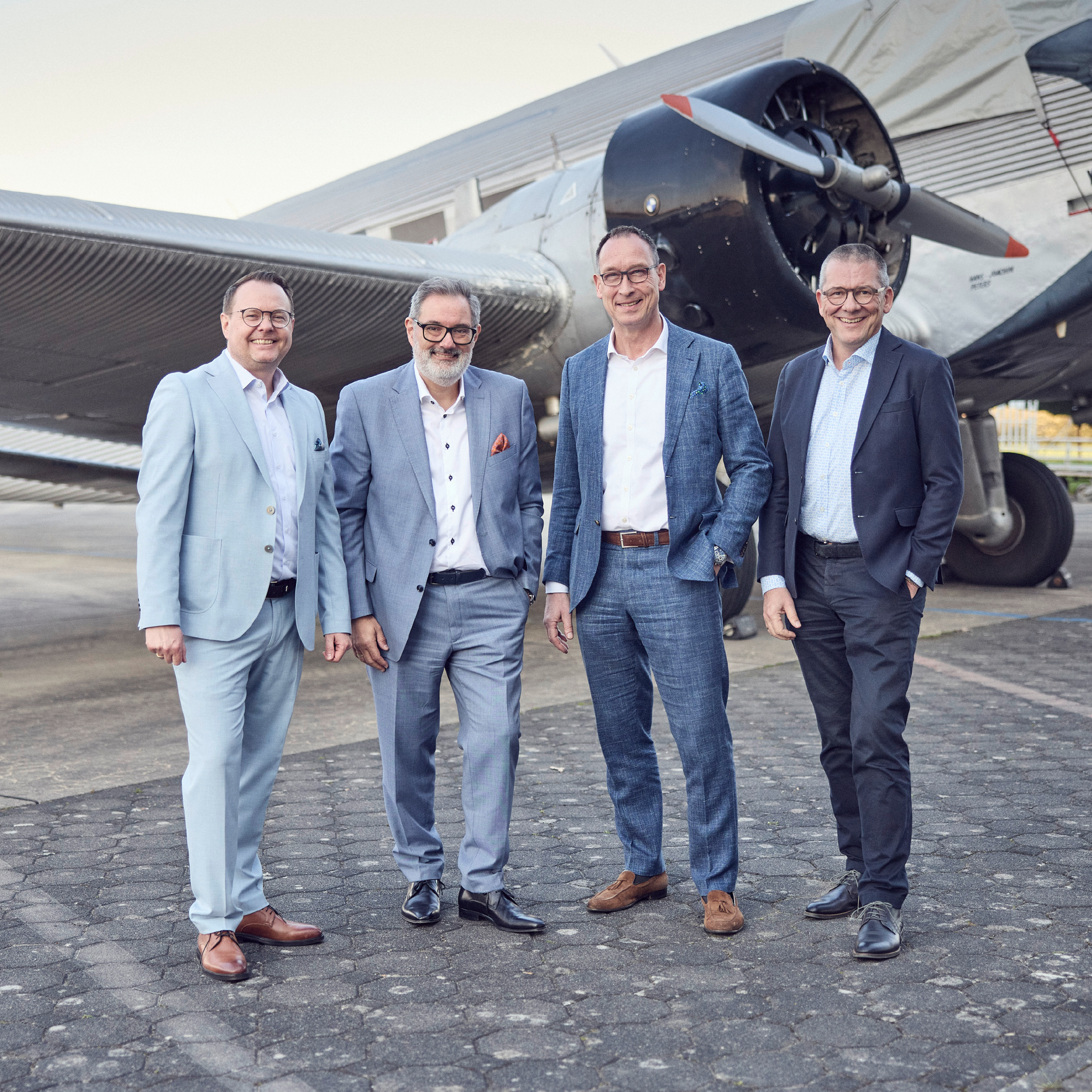 Vier Männer in formeller Kleidung stehen lächelnd vor einem historischen Flugzeug auf einem Flughafen.