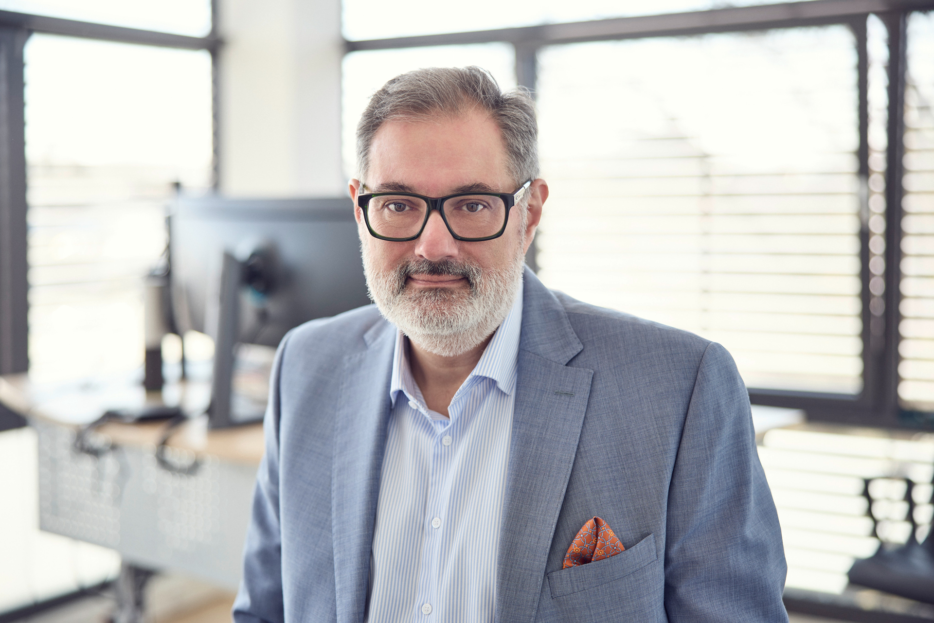 Ein Mann mit grauem Bart und Brille steht in einem modernen Büro, trägt einen hellblauen Anzug und ein gestreiftes Hemd. Im Hintergrund sind Büromöbel und große Fenster zu sehen, die Tageslicht hereinlassen.